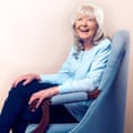 Jilly Cooper, in a long-sleeved shirt and with a fringe and shoulder-length hair, sits in an armchair and laughs
