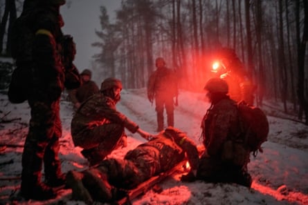 Men in army fatigues crouch or stand around a stretcher on the ground that appears to have a bundled-up man on it, in a dark forest in the snow