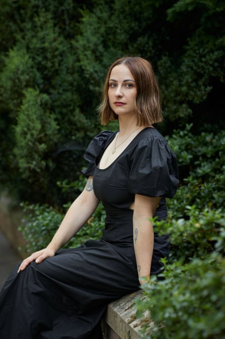 Lara Williams in a long black low-cut evening dress sitting on a wall with green foliage behind her