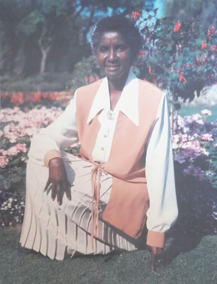 An old photo of an older black woman kneeling in front of flowers in a garden and smiling.