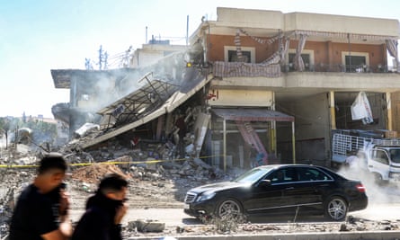 Two people cover faces as car passes partly destroyed building with dust and debris around