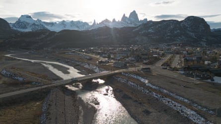 A road bridge over a river next to a small town at the foot of a mountain.