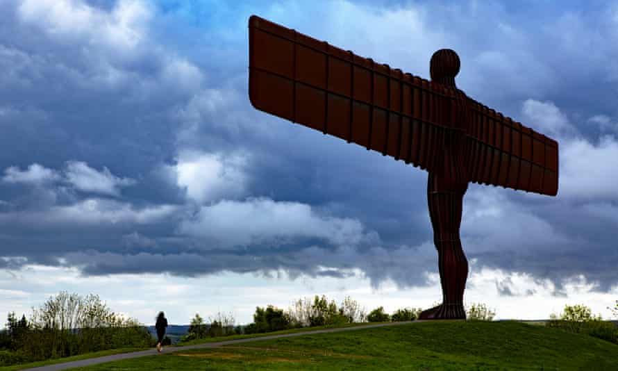 Angel of the North seen silhouetted from behind against a dramatic cloudy sky, with a jogger running on the road below