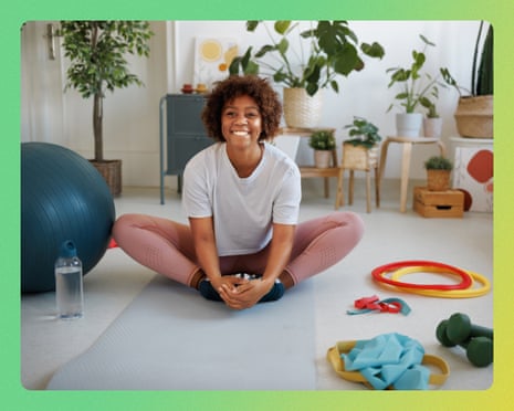 A smiling woman sits cross-legged on a yoga mat, warming up amid exercise equipment and plants