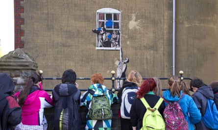Seven young people in winter jackets and backpacks look at a paintball marks on the wall graffittied with Banksy’s Well Hung Lover – a naked man hanging from a windowsill with a woman in a bra and pants and a man in a suit looking out from the window above him