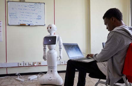 A young African man with a laptop sits in front of a small humanoid robot