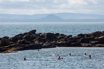 Swimmers in a tidal pool, seaparated from the sea by a band of rocks, with mountains in the distance
