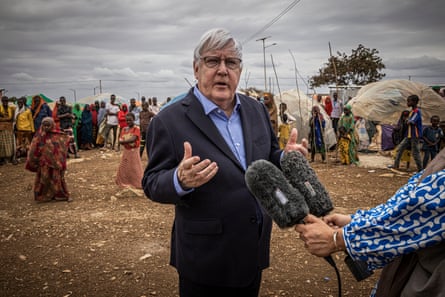 Martin Griffiths speaks into microphones held by a journalist as people at a displacement camp in Somalia stand and walk behind him