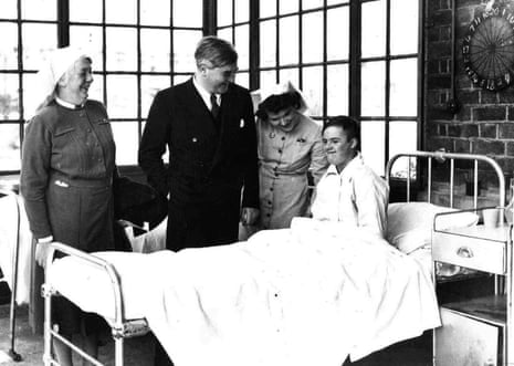 A black and white photo of Aneurin Bevan standing by a metal bed in an NHS hospital, with two nurses next to him and a boy sitting up in the bed