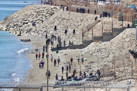 Dozens of people walk along a stony beach lined with security fencing