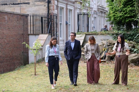 Andy Burnham stands in a grassy garden area with three women as one speaks as if she is explaining something. There are garden tools behind them and a brick wall by a large and grand-looking brick and stone building behind black iron railings.