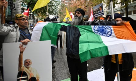 Amid a crowd of mostly Indian men, a man with a long black-and-white beard wearing a black skullcap pulls a green, white and orange flag in two.