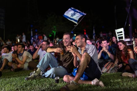 Young Israelis sitting on grass at night with an Israeli flag waving above them