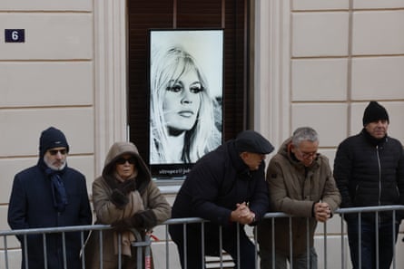 People dressed in warm jackets, some wearing hats and hoods, stand and lean on a metal barrier in a street. A large photo of the young Bardot is in a window behind them.
