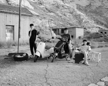 One boy reclines on a chair outside while two young scooterists stand by their vehicles