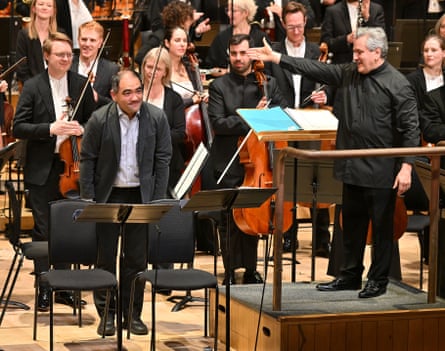 Conductor Sir Antonio Pappano gestures while smiling at Dai Fujikura standing among orchestra musicians