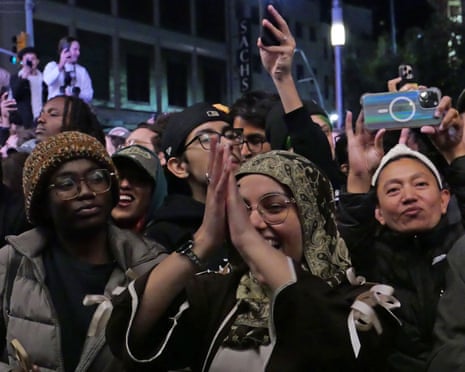 A woman in a headscarf claps her hand in celebration with other Mamdani supporters in a crowd.
