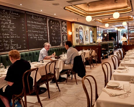 Diners sit at white-clothed tables in a restaurant with green banquettes, chalkboard menus, and decorative ceiling panels