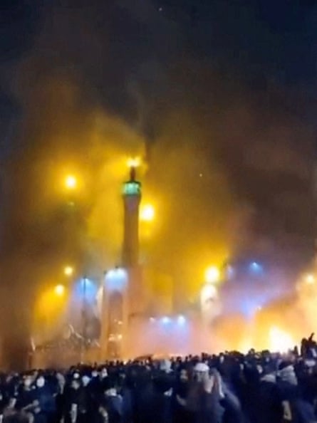 A mosque, lit up against the night sky, is surrounded by smoke with hundreds of protesters on the streets below