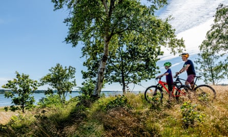 A Danish father and son standing with their bikes on a summer day amidst trees on the island of Mors in Denmark with the Limfjord in the background.