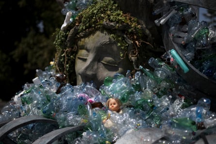 A sculpted head pokes out of a sea of plastic bottles and waste including a toy doll