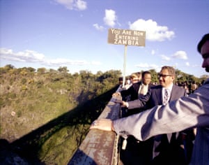 Kissinger and other black and white officials standing on a bridge with a sign in the background saying 'you are now entering Zambia'