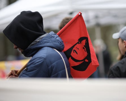 A man wearing a hoodie carrying a red flag bearing an image of Daniela Klette over his shoulder.