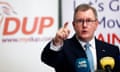 Jeffrey Donaldson speaks to the media during a press conference: he is pointing forwards and is seen in front of a DUP banner, and behind microphones. He wears a suit, tie and glasses.