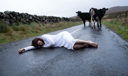 Ruth Wilson, barefoot and in a white nightgown, lies on a wet country road with two cows standing nearby