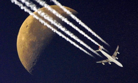 A plane flies past the half moon, leaving its contrails in the clear skies