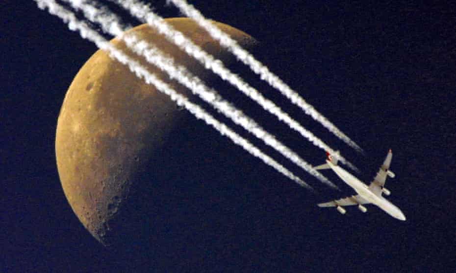 A plane flies past the half moon, leaving its contrails in the clear skies
