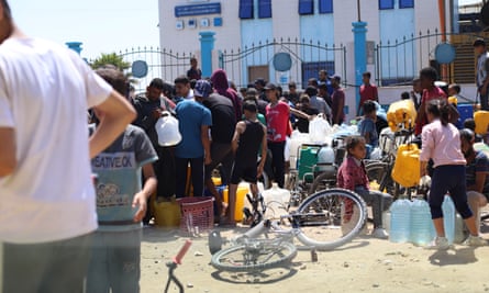 People stand and sit with large empty water bottles at the gates of a building