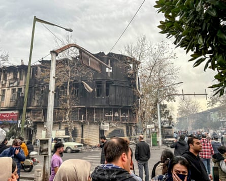 A street intersection full of people, many holding clothing to their faces, while in the background a charred building can be seen.