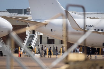 People line up near an aircraft behind a chain-link fence.