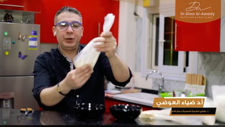 A video still of a man with safety goggles in a kitchen holding a plastic bag. In the corners of the screen are Arabic writing and in English his name above ‘nutrition and pain clinic’.