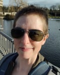 Anne Corwin, com cabelo muito curto e óculos de sol, sorri com um lago atrás dela