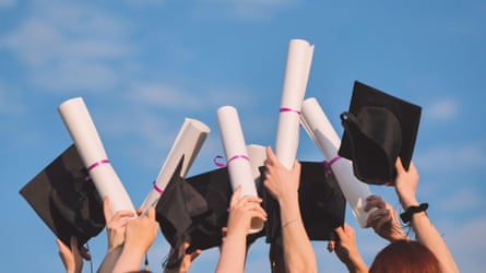 Graduates clasp up their diplomas and caps against a bluish sky.
