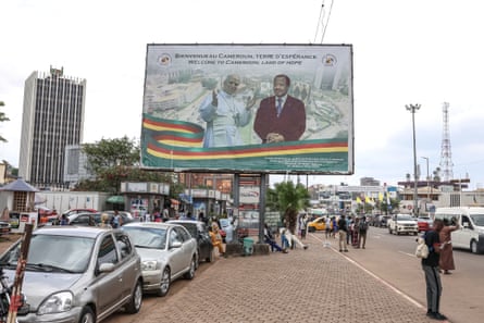 Cars and people beneath the billboard displaying portraits of the pair and a slogan saying ‘Welcome to Cameroon, land of hope’
