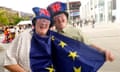 British Eurovision fans arrive at the venue for the grand final in Malmo, Sweden, 11 May 2024: two men laughing and wearing union jack-patterned sunglasses and hats, and one with a blue EU flag around his neck as well; the other man is holding this out to display the yellow stars. They are standing in the square in front of the concert hall.