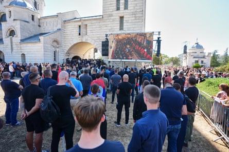 A crowd of people watch the funeral service on a large screen in front of a white stone Serbian Orthodox church with blue domes.