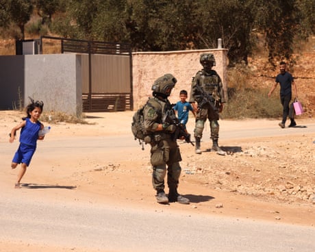 Children run through the street next to IDF soldiers in full combat gear