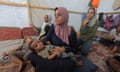 Infant held in his mother's arms; family members including three young girls are seen in the background as they sit on a rug and cushions inside a tent in a refugee camp