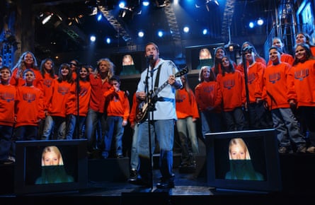 Adam Sandler appears on stage, playing a guitar in front of a choir of girls and boys in red sweatshirts with menorahs on them.