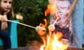 Two children hold marshmallows on sticks over a fire, one helped by an adult