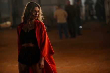 A young woman with long brown hair and wearing a red silk robe stands outdoors at night, with red dirt and figures in the background.