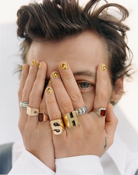 A closeup of Harry Styles with his hands covering most of his face, with lots of rings on and smiley faces on his fingernails