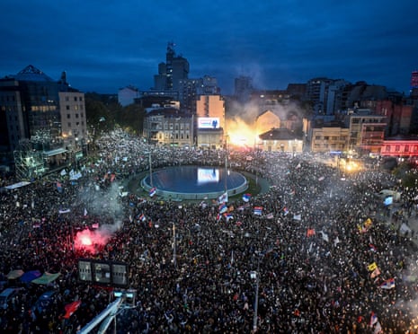 A huge crowd in Belgrade fills a square at night, holding phone lights and flags, near flares and smoke