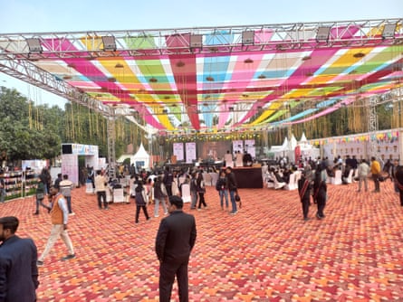 People mill about at an open air event space partly covered with multicoloured streamers with colourful flooring. At one end is a stage and some seating.