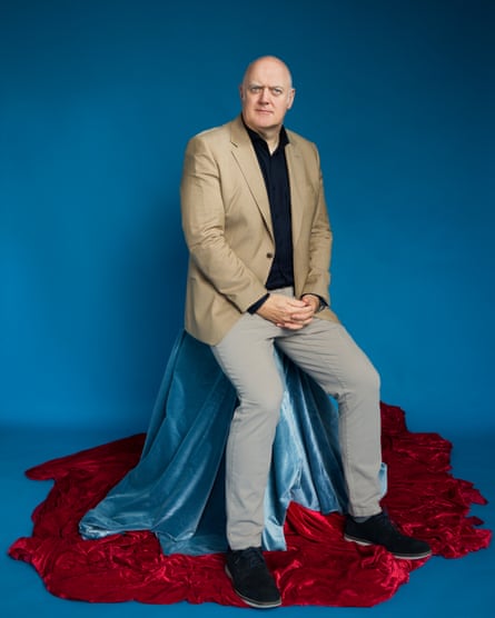 He sits in a photo studio atop a stool covered in a blue satin sheet, with a red one surround it at his feet