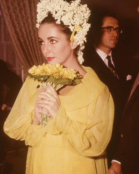 Taylor, wearing a yellow dress, holds a bouquet of yellow flowers and wears a headdress of white flowers